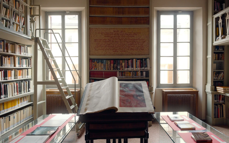 An open historic volume between the sunlit shelves of the Biblioteca di Busseto